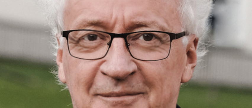 Ein Mann mit weißem Haar, der eine Brille und eine schwarze Jacke trägt, lächelt und schaut nach vorne.