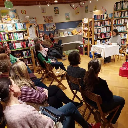Eine Frau liest aus einem Buch an einem Tisch in einer Bibliothek. Viele Menschen sitzen vor ihr in Stühlen.