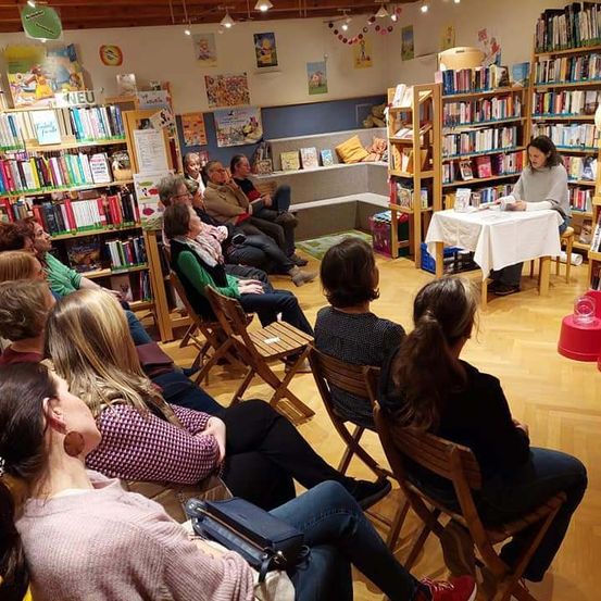 Eine Frau liest aus einem Buch an einem Tisch in einer Bibliothek. Viele Menschen sitzen vor ihr in Stühlen.
