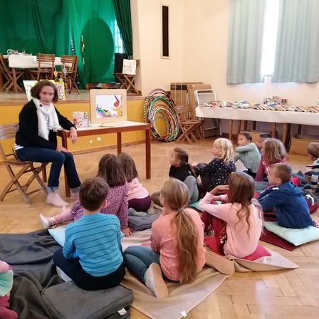 Eine Frau liest Kindern auf Kissen sitzend auf dem Boden ein Buch vor. Der Raum hat eine Bühne mit grünen Vorhängen und einem Tisch mit Büchern. Stühle und ein langer Tisch sind im Hintergrund.