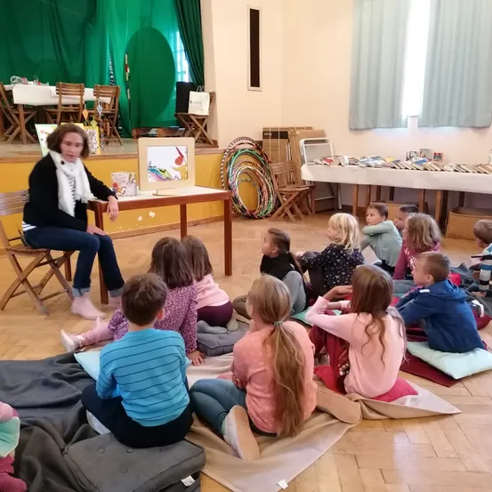 Eine Frau liest Kindern auf Kissen sitzend auf dem Boden ein Buch vor. Der Raum hat eine Bühne mit grünen Vorhängen und einem Tisch mit Büchern. Stühle und ein langer Tisch sind im Hintergrund.