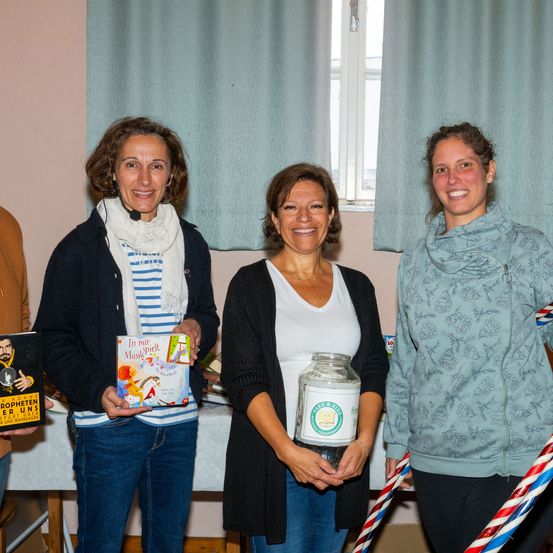 Drei Frauen stehen nebeneinander und halten ein Buch, ein Glas und ein Band. Sie lächeln und scheinen für ein Foto zu posieren. Die Frau auf der linken Seite hält ein Buch mit dem Titel 'In mir Musik spielt'. Die Frau in der Mitte hält ein Glas mit einem Etikett, das 'Park & Ride' besagt. Die Frau auf der rechten Seite hält ein Band. Hinter ihnen ist ein Fenster mit Vorhängen.