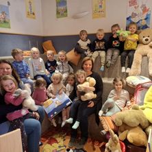 Eine Gruppe von Kindern und zwei Frauen sitzt auf einer Couch und hält Plüschtiere. Eine Frau hält ein Buch und ein kleines Mädchen. Eine Wand mit Bilderrahmen und einer Lampe.