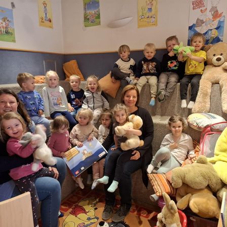 Eine Gruppe von Kindern und zwei Frauen sitzt auf einer Couch und hält Plüschtiere. Eine Frau hält ein Buch und ein kleines Mädchen. Eine Wand mit Bilderrahmen und einer Lampe.