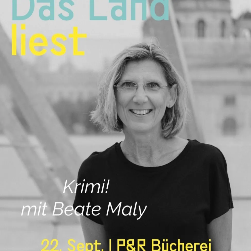 Ein Schwarz-Weiß-Foto einer lächelnden Frau mit Brille. Sie steht vor einem Gebäude mit dem Text 'Das Land liest' und 'Krimi! mit Beate Maly'. Das Foto wurde am 22. September in der LP&R Bucherei aufgenommen.