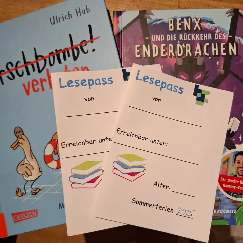 Zwei weiße Karten mit dem Text 'Lesepass' und 'Erreichbar unter' liegen auf drei Büchern. Die Bücher haben bunte Cover und Titel wie 'Ulrich Hub', 'BENX - und die Ruckkehr des Enderdrachen' und 'Schbombel'. Die Karten haben Illustrationen von Büchern und einer Ente.