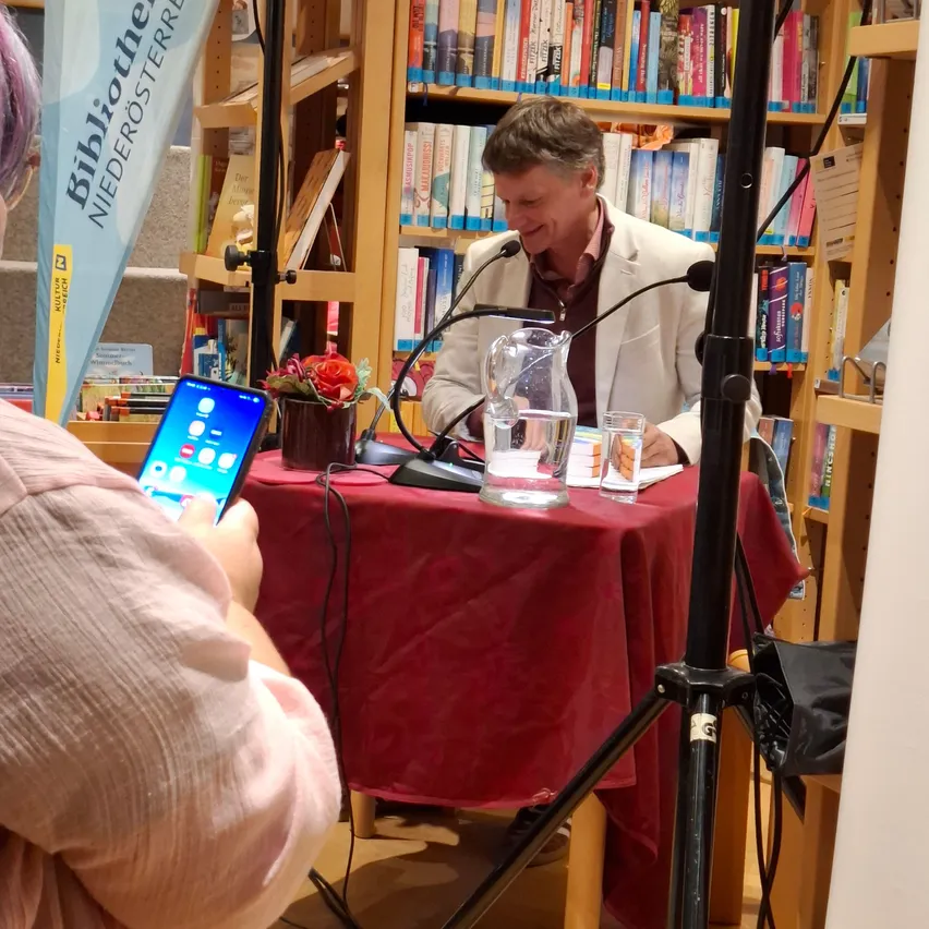 Ein Mann spricht in ein Mikrofon an einem Tisch in einer Bibliothek, mit einem Telefon und Blumen im Vordergrund.