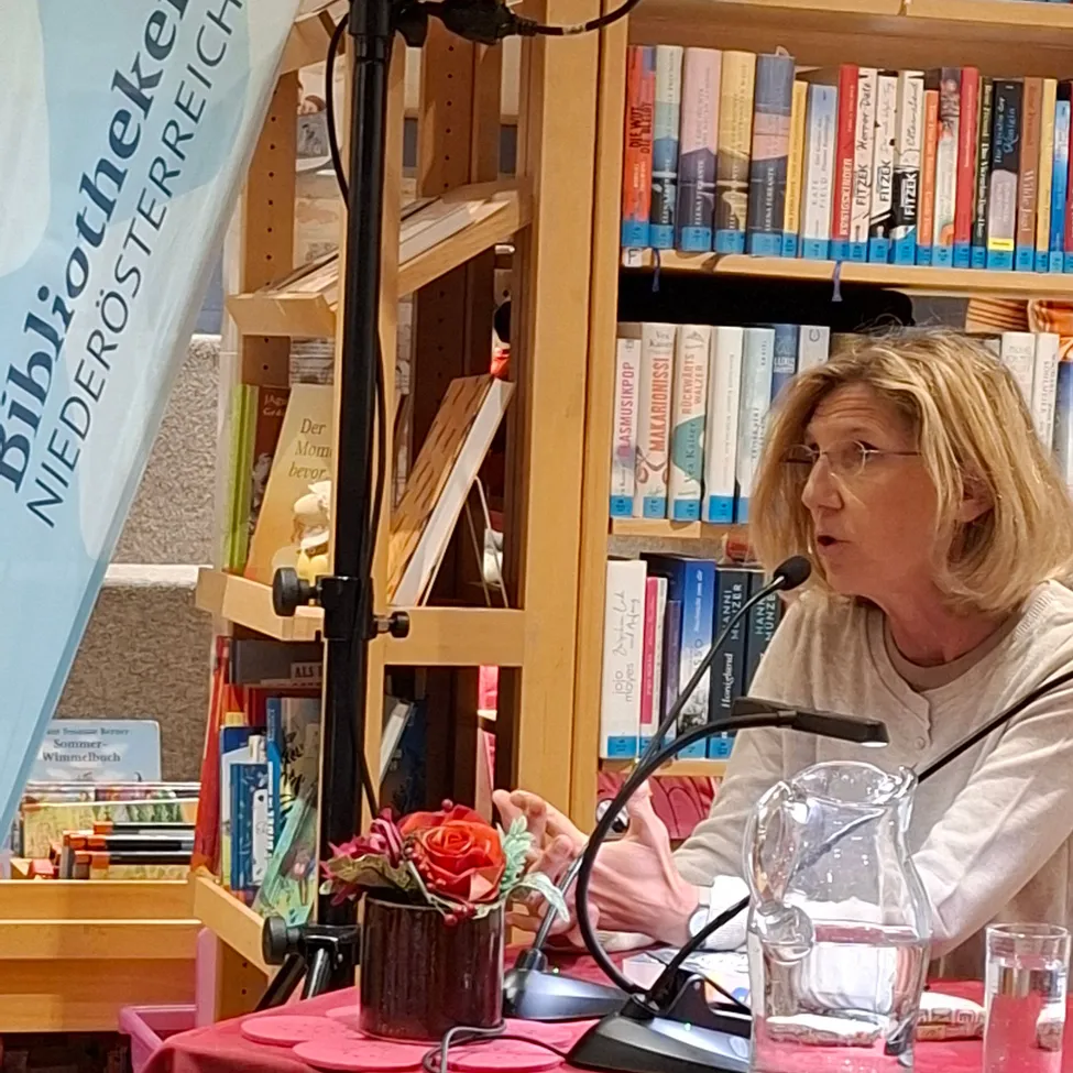 Eine Frau mit Brille spricht in ein Mikrofon an einem Rednerpult, mit einem Blumenvase und einem Glas Wasser auf dem Tisch vor ihr. Hinter ihr befinden sich mit Büchern gefüllte Bücherregale. Ein Banner mit dem Text 'Bibliothek Niederosterreich' ist links sichtbar.