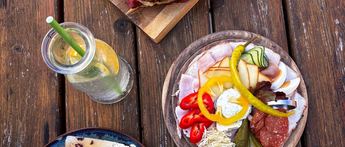 Auf einem Holztisch stehen ein Sandwich auf einem Schneidebrett, eine gemischte Gemüseplatte, ein Salat mit Eiern und Käse und ein Glas mit Strohhalm.