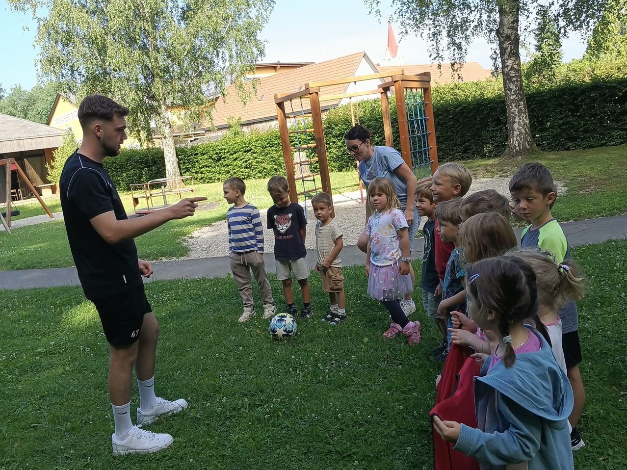 A group of children and adults are playing soccer in a park with a playground in the background.