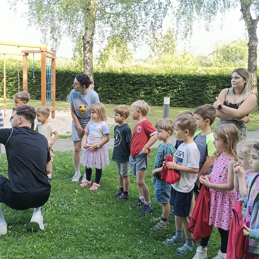 Several children stand on grass near a playground with adults, one wearing glasses and another a watch. Trees and hedges are in the background.
