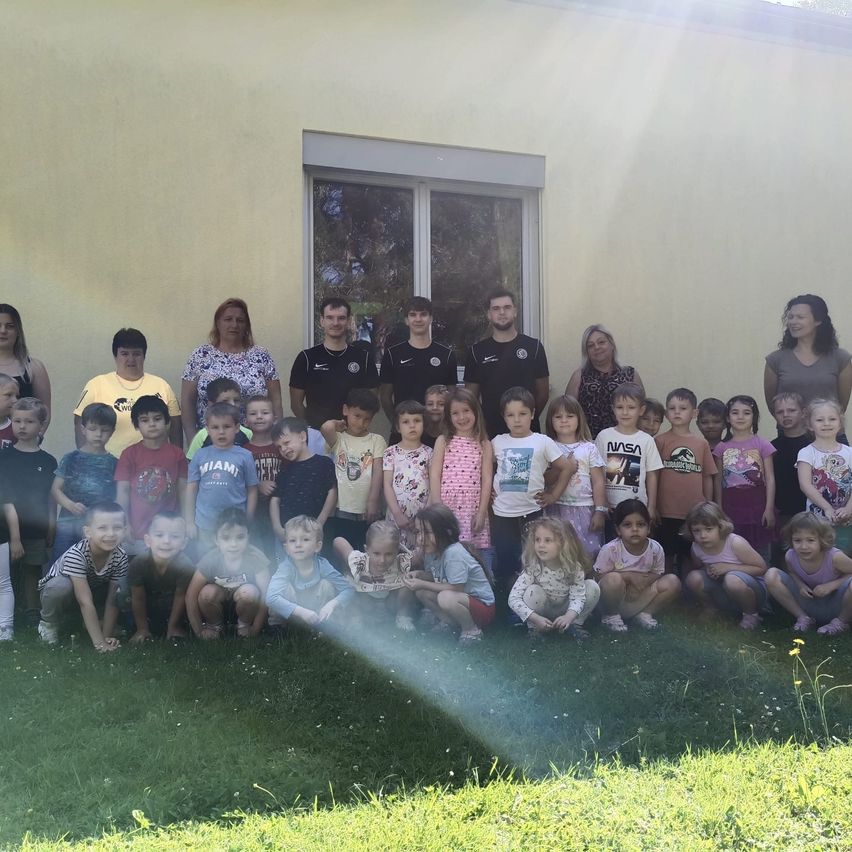 A group of children and adults pose for a photo in front of a yellow building. Some wear t-shirts with graphics, while others wear sneakers. The adults stand in the back.