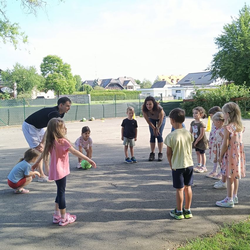 Several children are gathered in a park with two adults. One adult is bending over, and the other is standing. The children are in different positions, some standing and some sitting. Trees and houses are in the background.