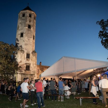 Eine Gruppe von Menschen versammelt sich bei Dämmerung vor einem alten Gebäude unter einem Zelt. Ein hoher, verwitterter Turm steht im Hintergrund.