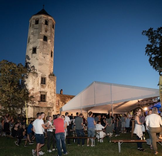 Eine Gruppe von Menschen versammelt sich bei Dämmerung vor einem alten Gebäude unter einem Zelt. Ein hoher, verwitterter Turm steht im Hintergrund.