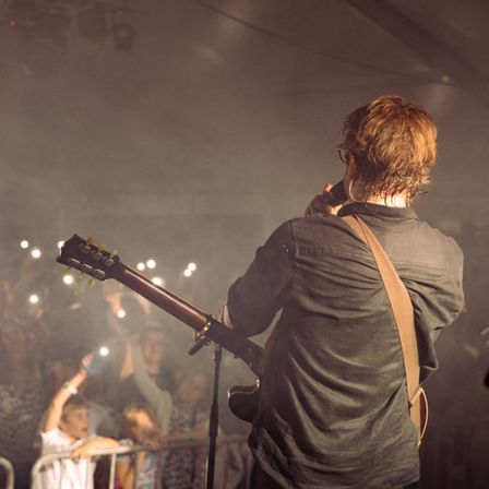 Ein Mann spielt Gitarre auf der Bühne bei einem Konzert, mit Blick auf das Publikum, während Lichter im Hintergrund blinken.