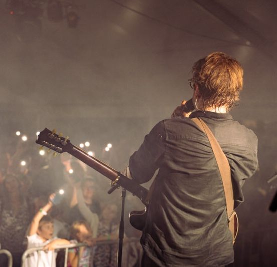 Ein Mann spielt Gitarre auf der Bühne bei einem Konzert, mit Blick auf das Publikum, während Lichter im Hintergrund blinken.