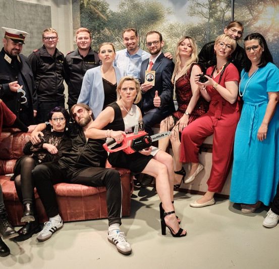 Eine Gruppe von Menschen in verschiedenen Kostümen posiert für ein Foto vor einer Kulisse mit einem Gemälde eines Waldes. Einige sitzen auf einer Couch, während andere stehen.