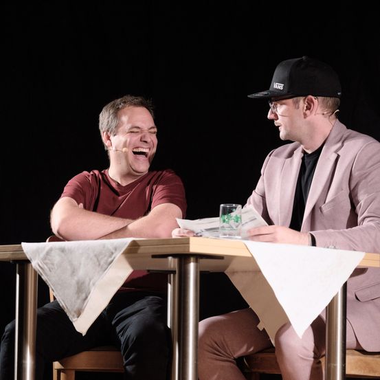 Zwei Männer sitzen an einem Tisch, einer lacht und der andere liest eine Zeitung. Der lachende Mann hat ein Mikrofon im Ohr, und der andere trägt einen schwarzen Hut und eine Brille.