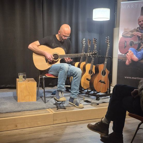Ein Mann spielt Gitarre auf einer Bühne mit mehreren Gitarren dahinter. Ein Poster mit einem Gitarre spielenden Mann ist an der Wand.