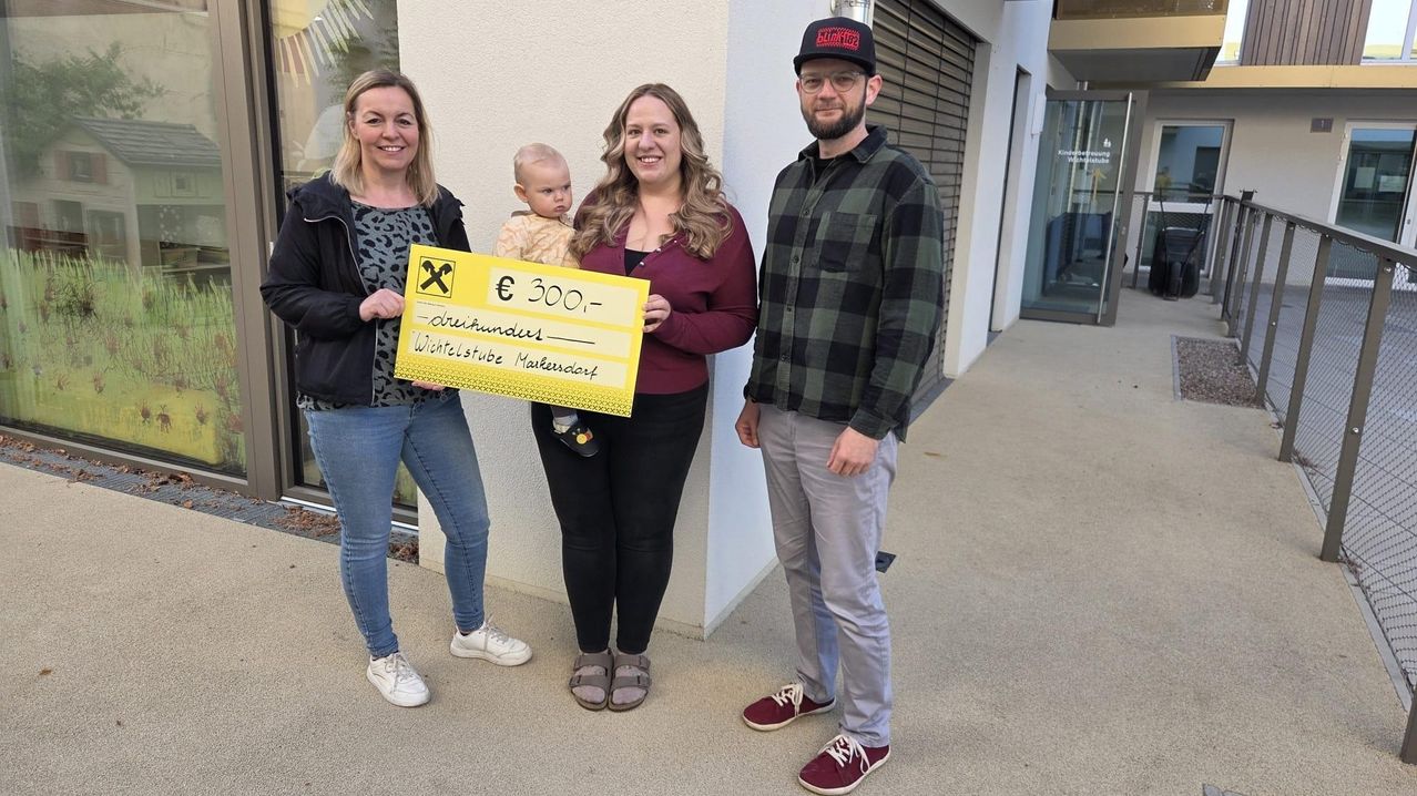 Drei Personen posieren für ein Foto mit einem gelben Scheck. Die Frau links hält ein Baby, die Frau in der Mitte hält einen Scheck und der Mann rechts trägt eine Kappe.
