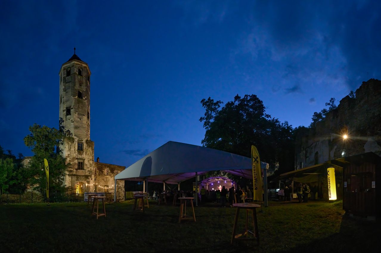 Ein großes Zelt vor einer alten Burg bei Nacht, mit Menschen im Inneren und einer Banner an der Seite.