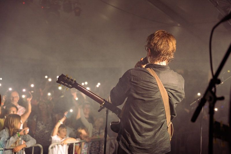 Ein Musiker spielt Gitarre auf der Bühne bei einem Konzert, mit einer Menge Menschen im Hintergrund.