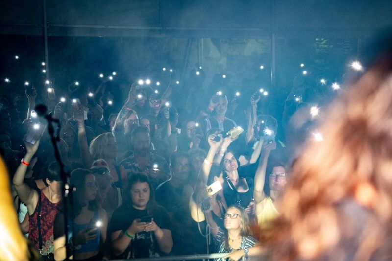 Eine Menschenmenge bei einem Konzert, viele halten ihre Telefone mit Taschenlampen hoch und erzeugen ein Meer leuchtender Lichter. Die Atmosphäre ist erfüllt von Rauch und Aufregung.