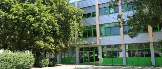 Ein modernes Bürogebäude mit grünem Rand ist von Bäumen und Büschen umgeben. Das Gebäude hat viele Fenster und eine grüne Tür.