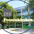 Ein blauer Kreis mit 'pts Frauenkirchen' in gelber Schrift um ein Bild eines modernen Gebäudes mit grünen Fenstern und einer von Bäumen gesäumten Vorderseite.