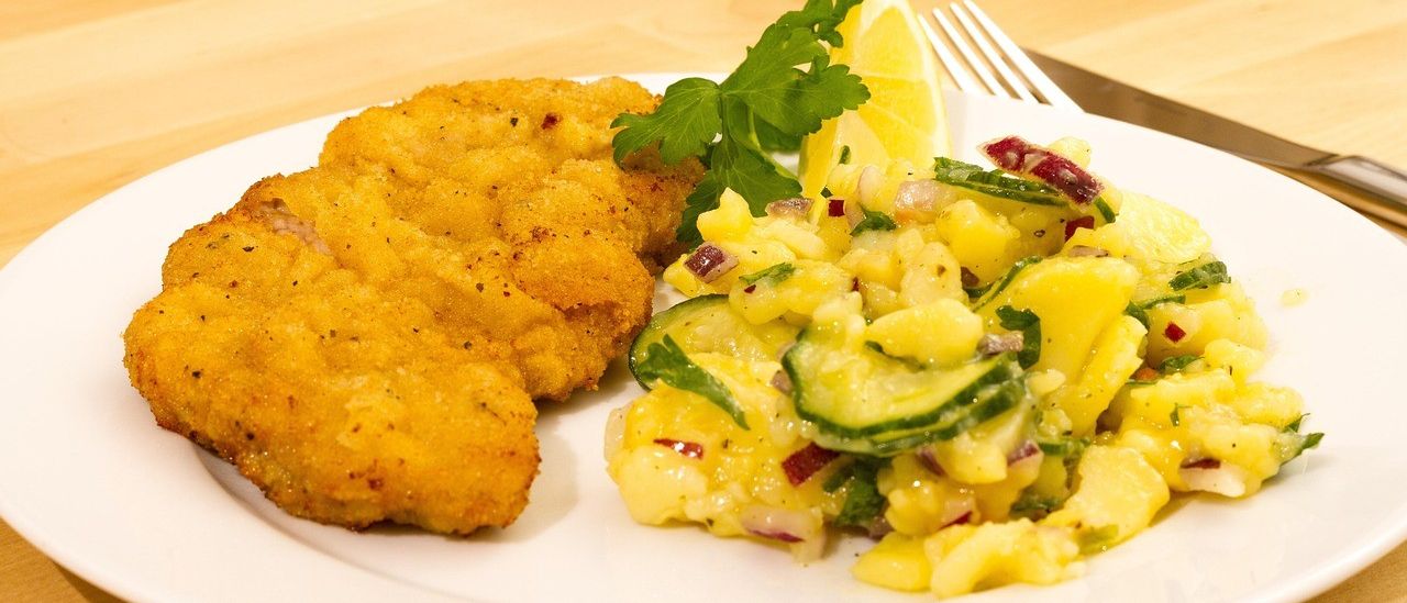 Ein Teller mit zwei Stücken panierter Chicken, einer Kartoffelsalat mit Gurke und einer Zitronenscheibe, auf einem Holztisch.