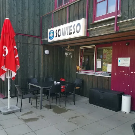 Eine Außensitzfläche eines Restaurants namens Sowieso. Der Bereich hat einen schwarzen Tisch und Stühle, einen roten Schirm und eine rote Tür.