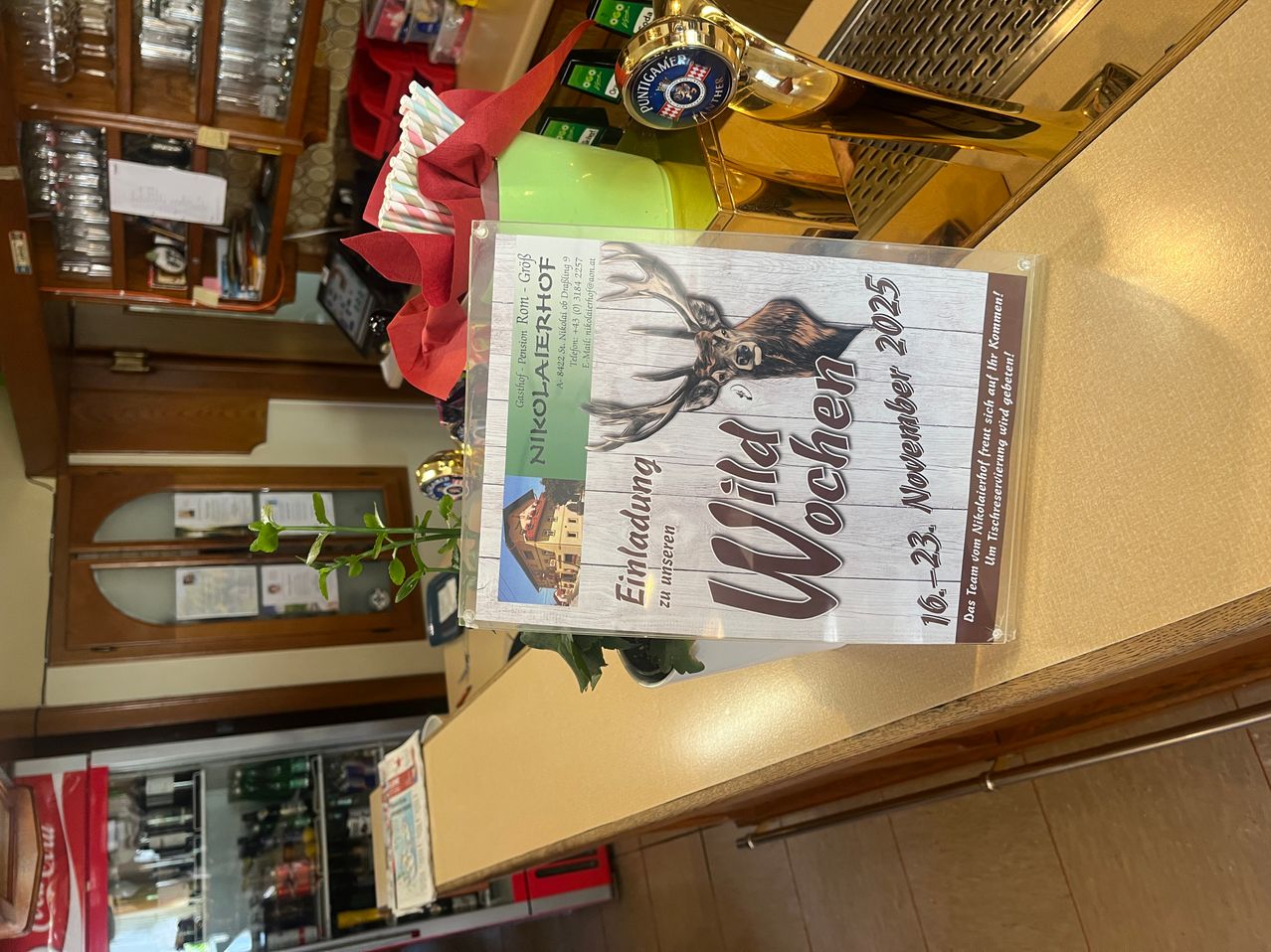 Ein durchsichtiges Kunststoffgehäuse zeigt ein Poster mit einem Hirschbild, der Text lautet 'Wildes Wochenende' und 'Nikolaterhof'. Das Poster ist an einer Holztheke befestigt. Dahinter sind verschiedene Gegenstände wie Flaschen, Gläser und ein Bierhahn zu sehen.