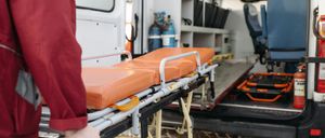 Eine Person in einem roten Uniform hält eine orange Trage aus einem Krankenwagen mit einer aufgeschobenen Tür.