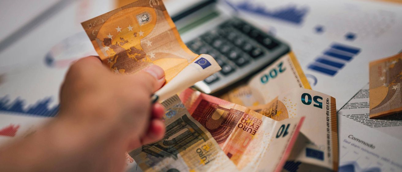 Eine Hand hält eine Euro-Banknote über einem Taschenrechner und einem Haufen Euro-Banknoten.