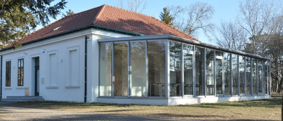 Ein weißes Gebäude mit rotem Dach und großen Glasfenstern, umgeben von kahlen Bäumen und einer Kiesauffahrt unter einem klaren Himmel.