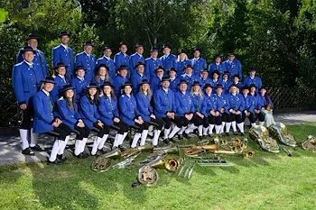 Eine Gruppe von Musikern in blauen Uniformen und schwarzen Hüten posiert für ein Foto mit ihren Instrumenten auf einem Rasenfeld.
