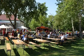 Ein großer Außenbereich mit mehreren Picknicktischen und Bänken. Menschen sitzen um die Tische, einige mit Getränken. Es gibt Bäume, weiße Sonnendächer und ein Gebäude im Hintergrund.