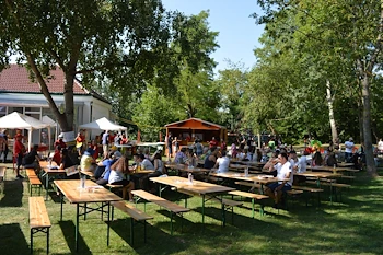 Ein großer Außenbereich mit mehreren Picknicktischen und Bänken. Menschen sitzen um die Tische, einige mit Getränken. Es gibt Bäume, weiße Sonnendächer und ein Gebäude im Hintergrund.