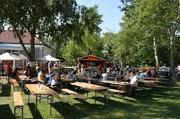 Viele Menschen sitzen an hölzernen Picknicktischen in einem Park mit grünem Rasen, Bäumen und einem Gebäude mit rotem Dach. Im Hintergrund befinden sich weiße Vordächer und ein kleines orangefarbenes Gebäude.