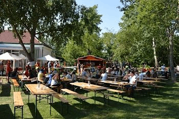 Viele Menschen sitzen an hölzernen Picknicktischen in einem Park mit grünem Rasen, Bäumen und einem Gebäude mit rotem Dach. Im Hintergrund befinden sich weiße Vordächer und ein kleines orangefarbenes Gebäude.