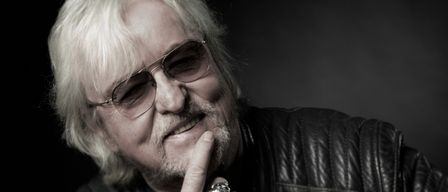 Ein Schwarz-Weiß-Porträt eines älteren Mannes mit Sonnenbrille, Lederjacke und Ring, mit einem Finger auf den Lippen.
