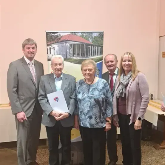 Fünf Personen stehen eng beieinander für ein Foto. Ein Mann hält ein Papier mit einem Logo. Dahinter ist eine Wand mit einem Hausposter. Ein Tisch mit einer weißen Tischdecke steht rechts.