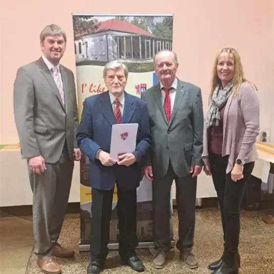 Vier Personen stehen vor einem Hausbanner. Der Mann in der Mitte hält ein Papier.