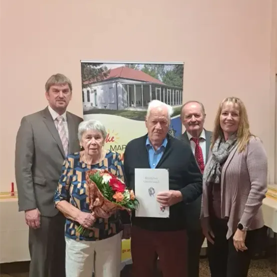 Fünf Personen stehen in einem Raum, ein älterer Mann hält ein Papier mit einem Logo, eine ältere Frau hält einen Blumenstrauß und ein Banner ist hinter ihnen.