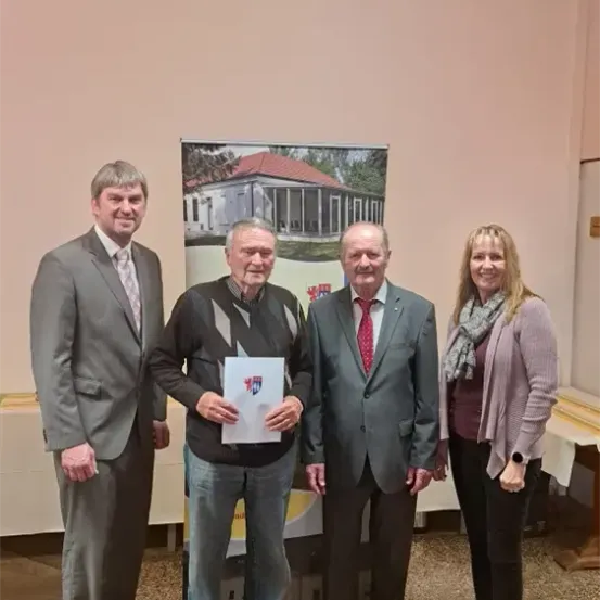 Vier Personen stehen in einem Raum zusammen. Der Mann in der Mitte hält ein Zertifikat. Hinter ihnen ist ein Banner mit einem Hausbild. Sie alle lächeln und scheinen für ein Foto zu posieren.