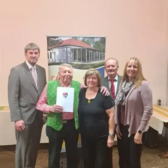Fünf Personen stehen zusammen vor einem Banner. Der Mann in der Mitte hält ein Stück Papier, und die beiden Frauen neben ihm lächeln. Der Mann links trägt einen Anzug mit Krawatte, während der Mann rechts eine rote Krawatte trägt. Hinter ihnen steht ein Tisch mit einer weißen Tischdecke.