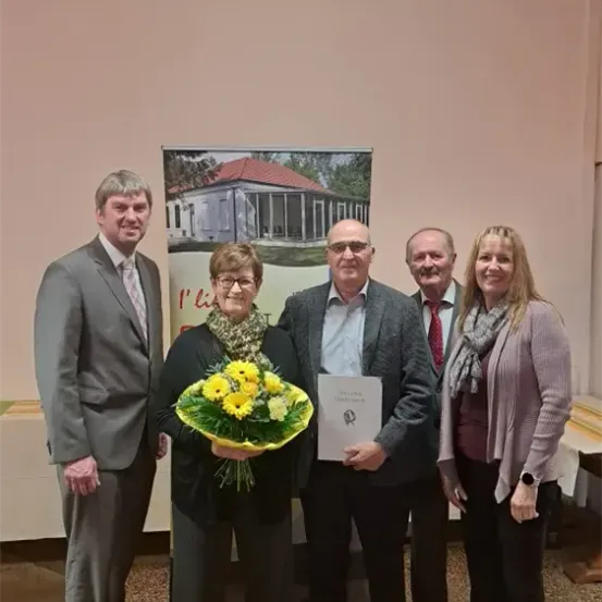 Fünf Menschen stehen lächelnd in einem Raum. Eine Frau hält einen Blumenstrauß und ein Zertifikat. Ein Banner mit einem Hausbild steht hinter ihnen.