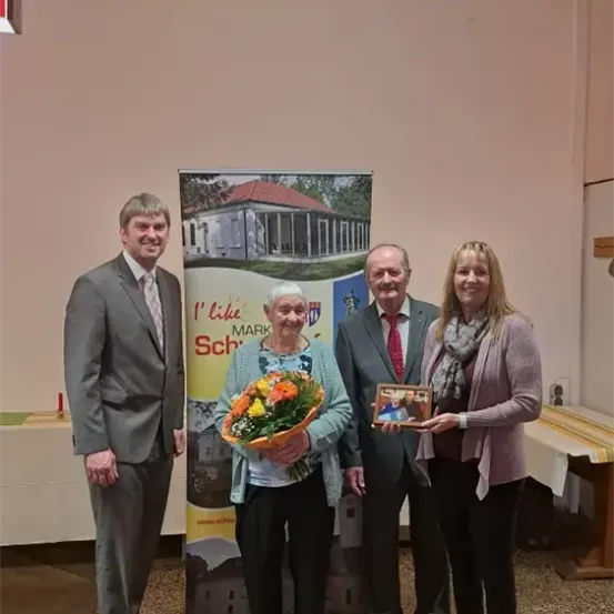 Vier Personen stehen vor einem Banner mit einem Bild eines Hauses. Die Frau hält Blumen und ein gerahmtes Bild. Der Mann links trägt einen Anzug und eine Krawatte.