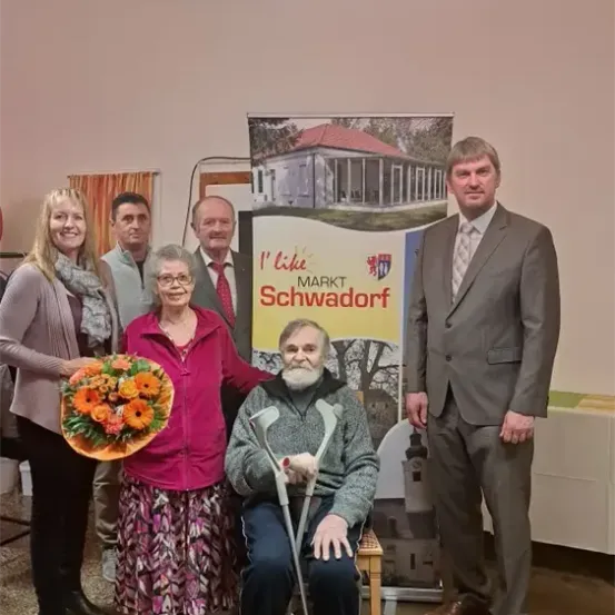 Eine Gruppe von Menschen, einschließlich eines älteren Mannes im Rollstuhl, versammelt sich für ein Foto. Eine Frau hält Blumen. Sie stehen vor einem Schild mit einem Hausbild und der Aufschrift 'Ich mag Markt Schwadorf'.